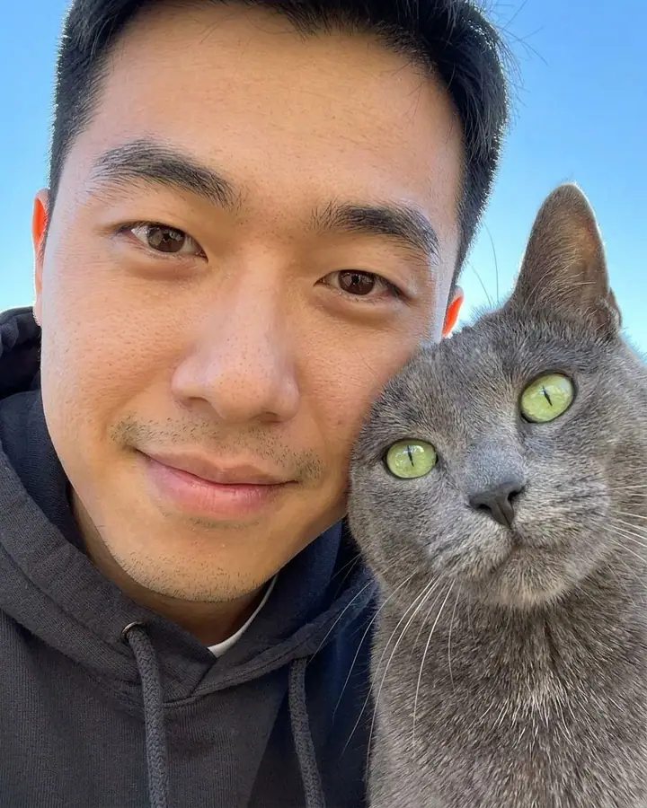 Young man taking a close-up selfie with a gray cat with green eyes
