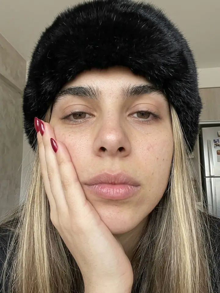 Very close front-camera selfie of a woman in a black fur hat resting her face on her hand